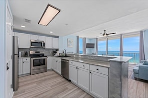 A modern kitchen with white cabinets, stainless steel appliances, and a large window overlooking the ocean.