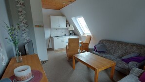 A cozy living room with a wooden ceiling, a dining table with a vase of flowers, a sofa with cushions, and a kitchen in the background with a skylight. - 2-zimmerwohnung in Direkter Strandnähe (Westerdeichstrich)
