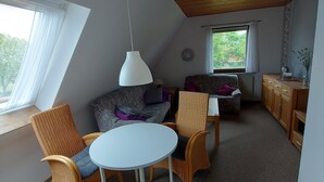 A cozy room with a sloped ceiling, a round table with chairs, a sofa with cushions, a small wooden cabinet, and a window with a view of trees. - 2-zimmerwohnung in Direkter Strandnähe (Westerdeichstrich)