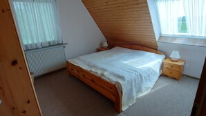 A bedroom with a sloped ceiling, a wooden bed, a nightstand, a radiator, and a window with sheer curtains. - 2-zimmerwohnung in Direkter Strandnähe (Westerdeichstrich)