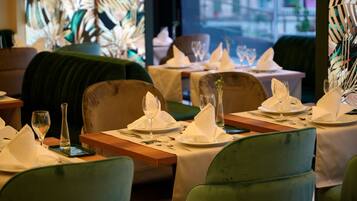 A restaurant interior with neatly set tables, green upholstered chairs, and a view of the street through large windows.