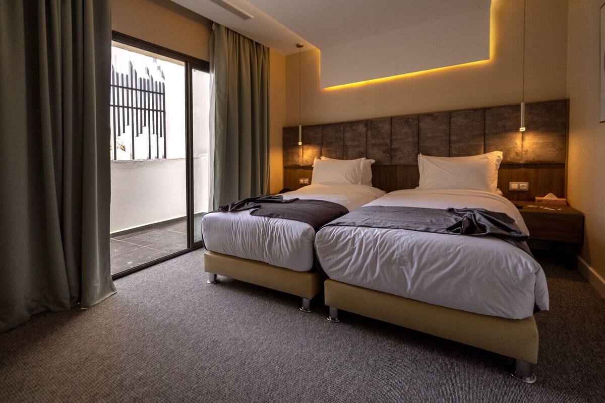 A hotel room with two beds, a large window with curtains, a headboard with built-in lighting, and a carpeted floor.
