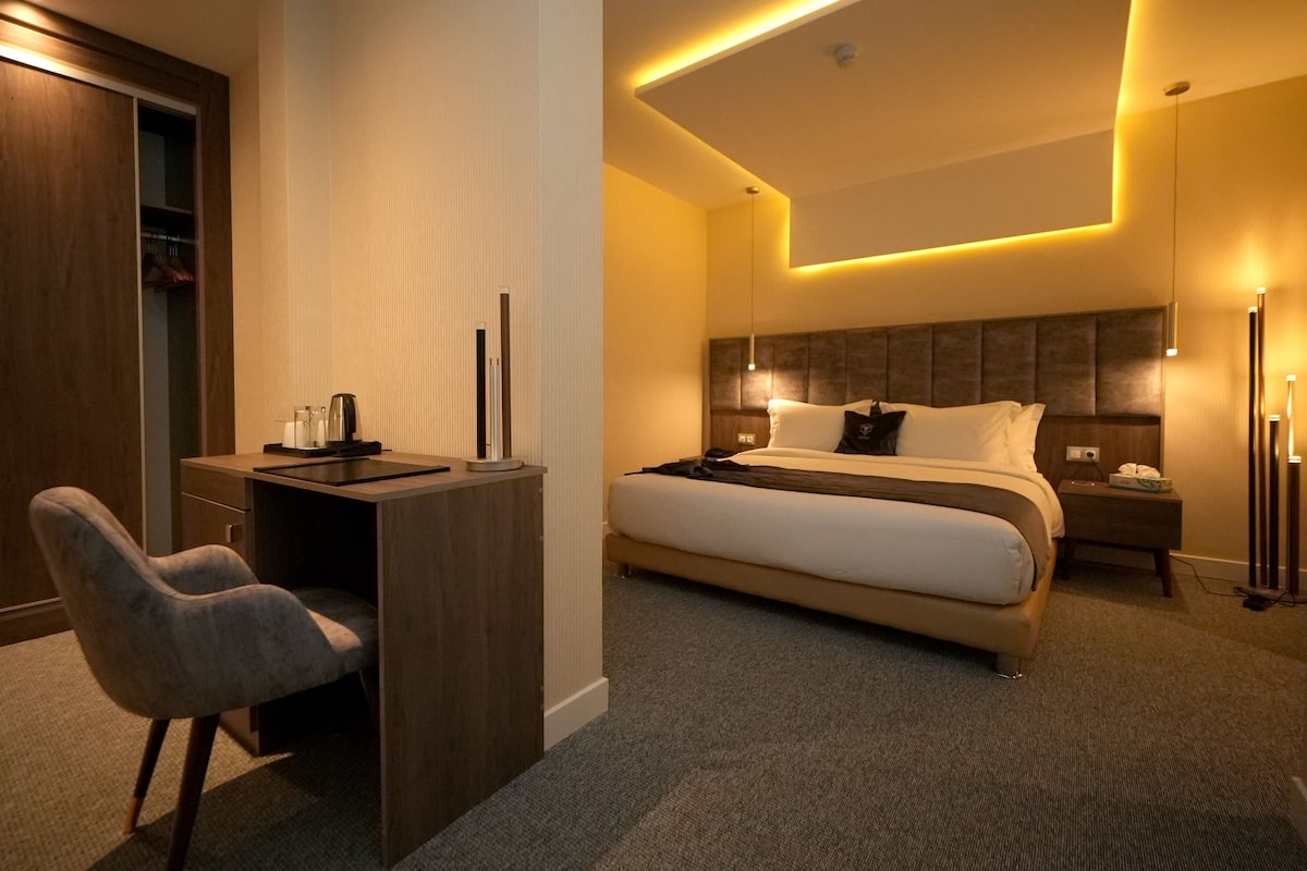 A modern hotel room with a bed, desk, chair, bedside table, and a wardrobe.