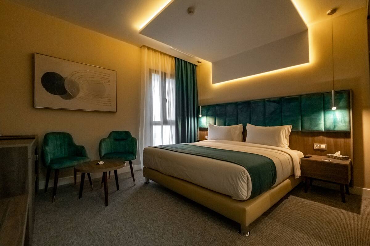 A modern hotel room with a large bed, two green armchairs, a small table, and a ceiling light fixture.