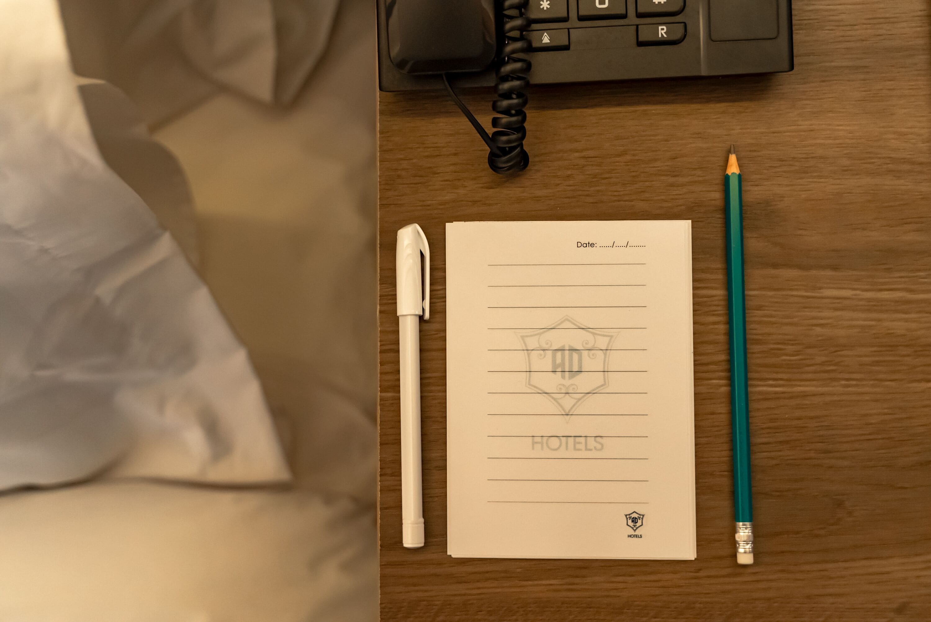 Een hotelkamer met een houten bureau, een telefoon, een notitieboekje met hotel logo, een pen en een potlood.