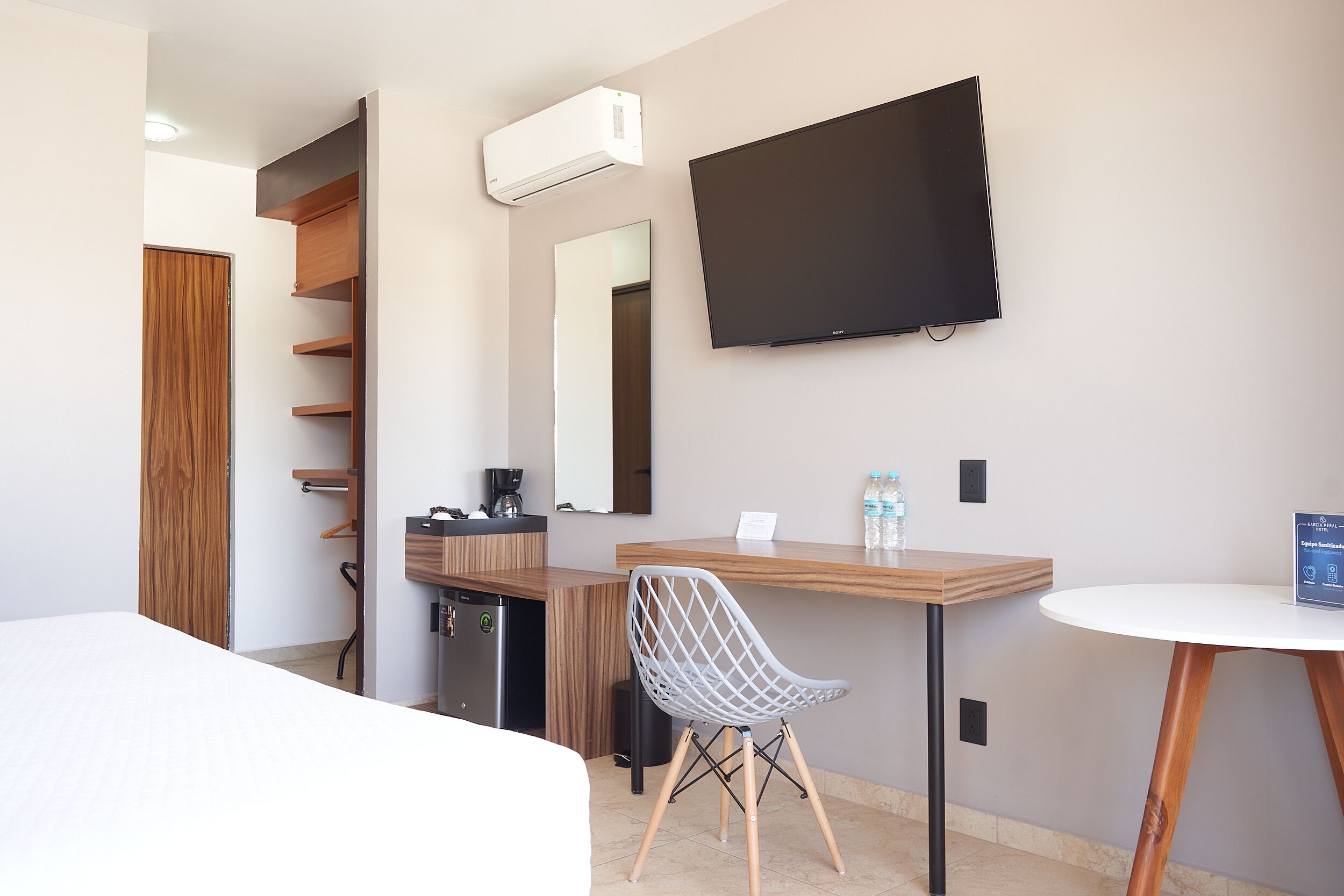 A hotel room with a bed, a desk with a chair, a TV, a mirror, a wardrobe, and a coffee machine.