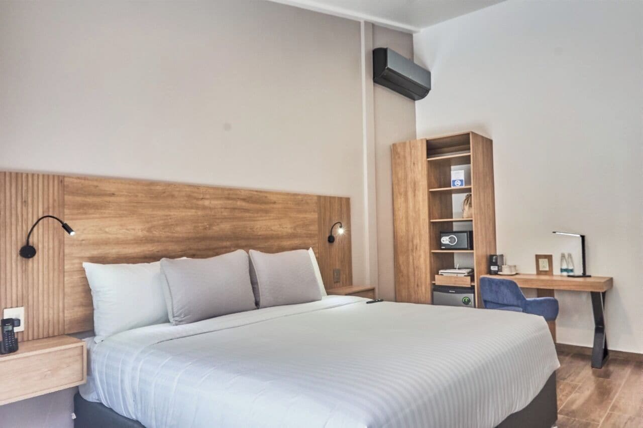 Una habitación de hotel moderna con una cama grande, cabecera de madera, un escritorio con silla, una estantería y un aire acondicionado de pared.