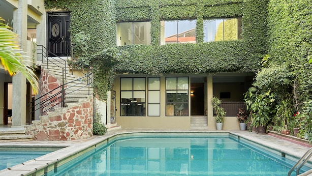Una piscina con borde de piedra, una escalera con barandilla metálica y un edificio parcialmente cubierto de vegetación.