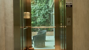 Un ascensor con puerta de vidrio, un cartel que indica "P1" y vista a un espacio exterior con vegetación y una silla de mimbre.