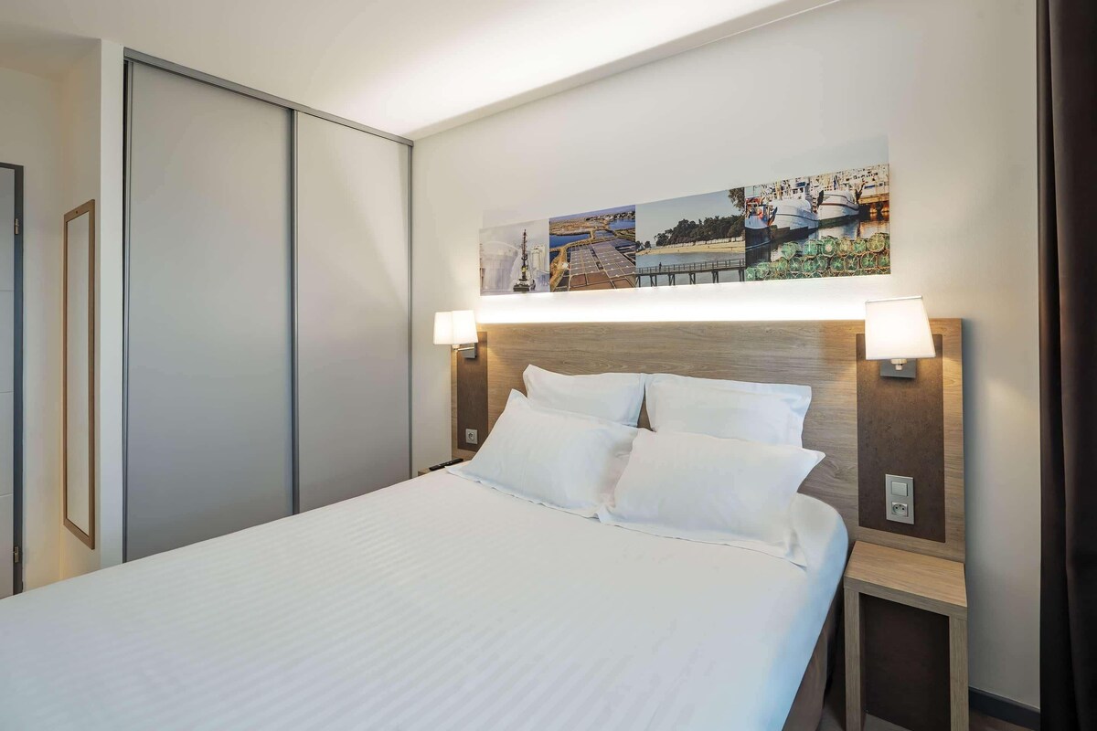 A hotel room with a bed, a nightstand, a wall-mounted lamp, and a painting above the bed.