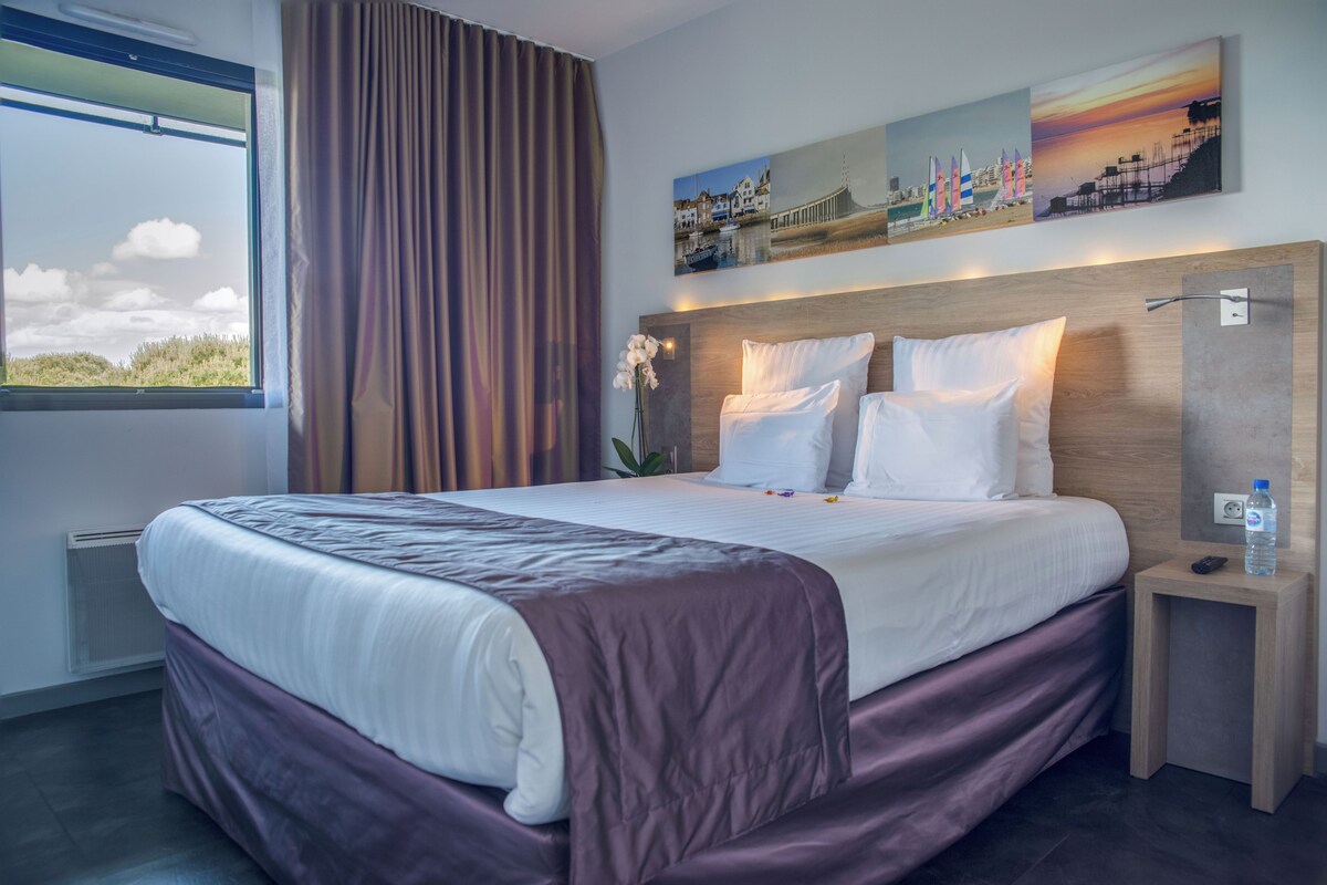 A hotel room with a large bed, a nightstand, a water bottle, and a painting on the wall.