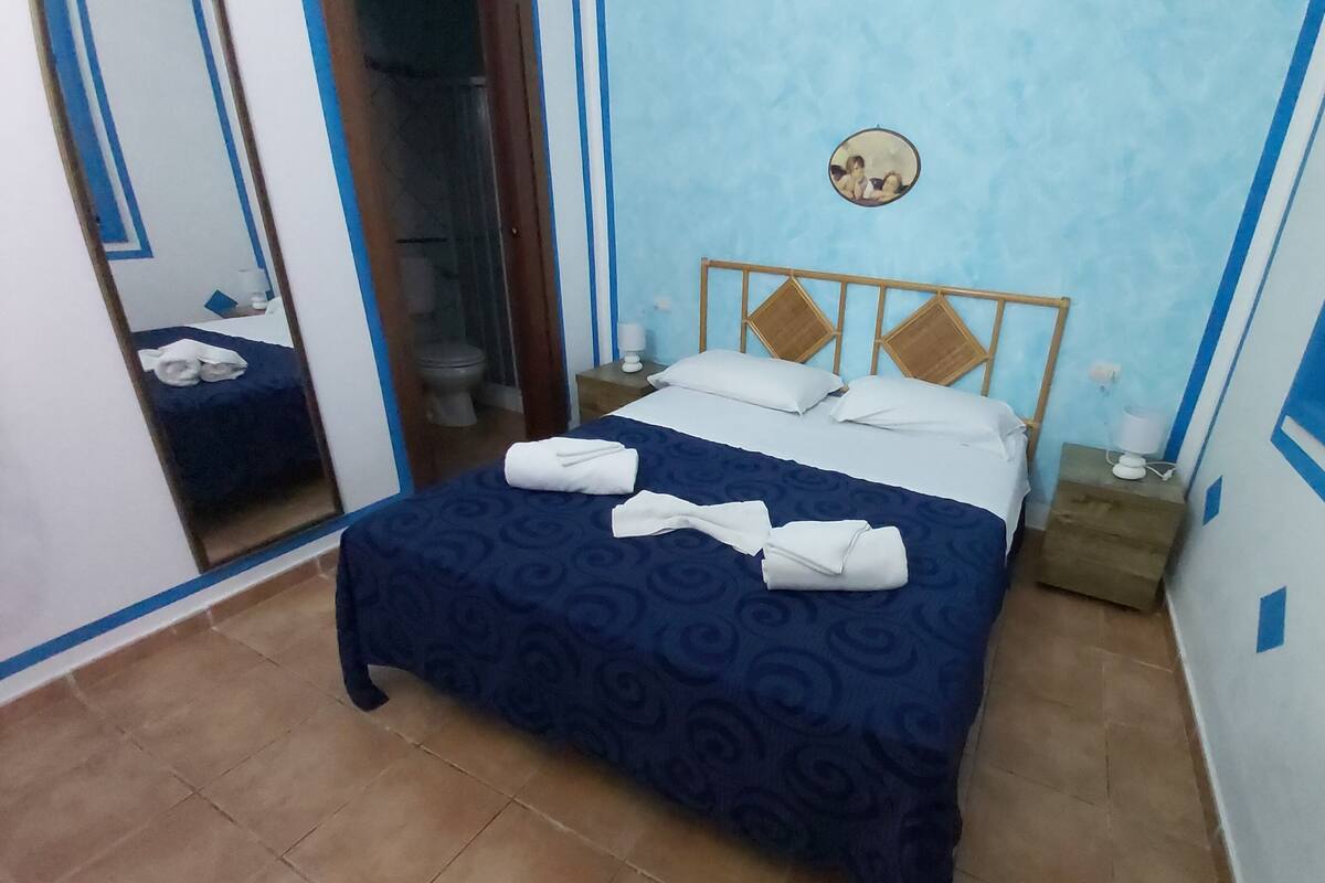 A hotel room with a bed, a mirror, a small bedside table, and a bathroom visible through a glass door.