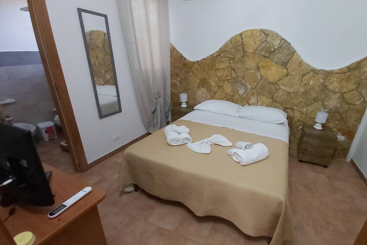 A hotel room with a stone headboard, a bed with white linens, a bedside table with a lamp, and a mirror on the wall.