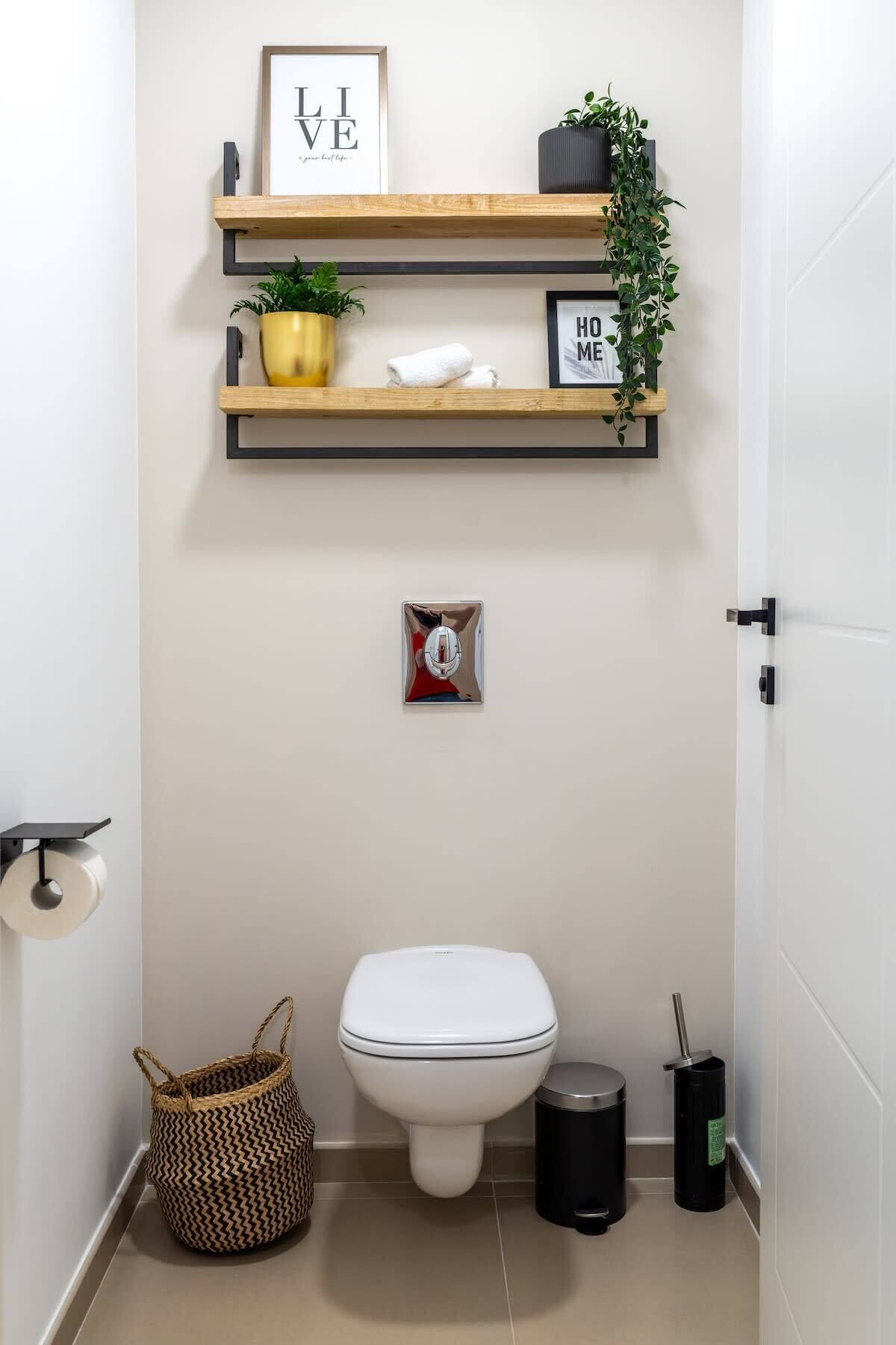 A small bathroom with a toilet, a trash bin, a basket, and two shelves with plants and framed items.