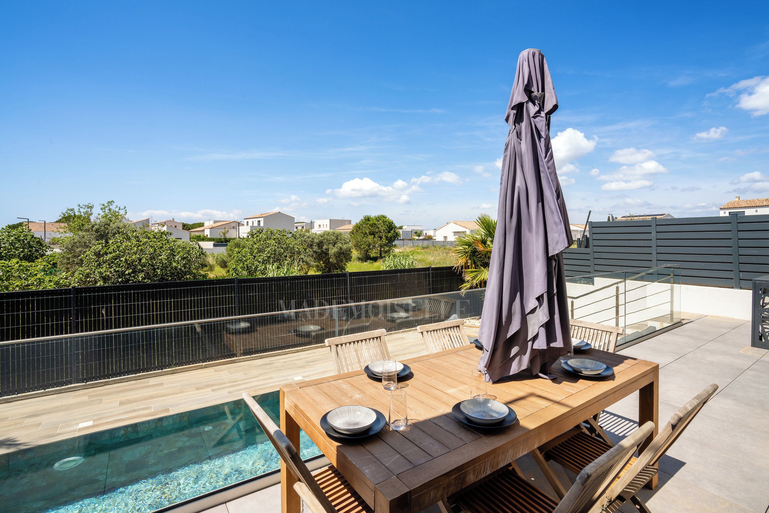 Terraço no telhado com piscina, mesa e cadeiras de madeira, com vista para uma área residencial.
