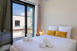 Une chambre d’hôtel avec un grand lit, deux oreillers, une table de chevet avec une lampe et une vue sur un immeuble à travers une porte vitrée.
