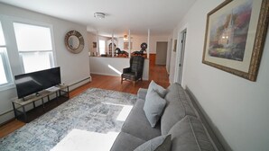 Un salon avec un canapé gris, un téléviseur à écran plat posé sur un meuble en bois, un tapis à motifs et un tableau accroché au mur.