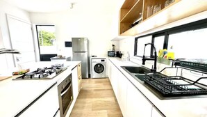 A modern kitchen with white cabinetry, a stainless steel stove, a built-in washing machine, and a window overlooking a green area. - Into The Sea - 5 mins from the Beach (Albion)