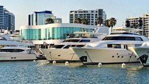 Marina con varios yates amarrados, rodeada de edificios modernos y palmeras.