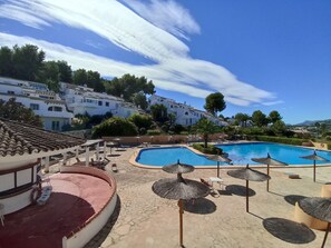 Un espace piscine d’un complexe hôtelier, avec un ciel d’un bleu limpide, des bâtiments blancs et des palmiers.