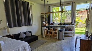 A modern hotel room with a bed, a sofa, a kitchen area with stools, and a view of the outdoors.