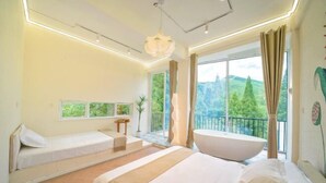 Un dormitorio moderno con una cama grande, una bañera integrada, un pequeño rincón de descanso y un balcón con vistas a zonas verdes.