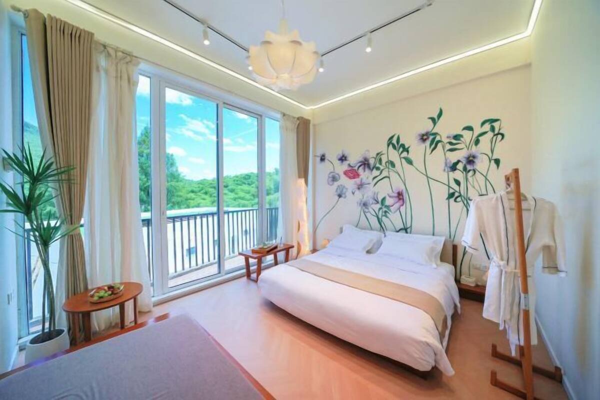 Un dormitorio con una cama grande, vistas a zonas verdes por la ventana, una mesita con una planta y un perchero.
