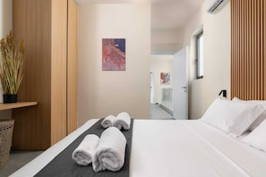 Quarto de hotel com cama, uma planta em vaso, um quadro na parede e um corredor que dá para outro quarto.