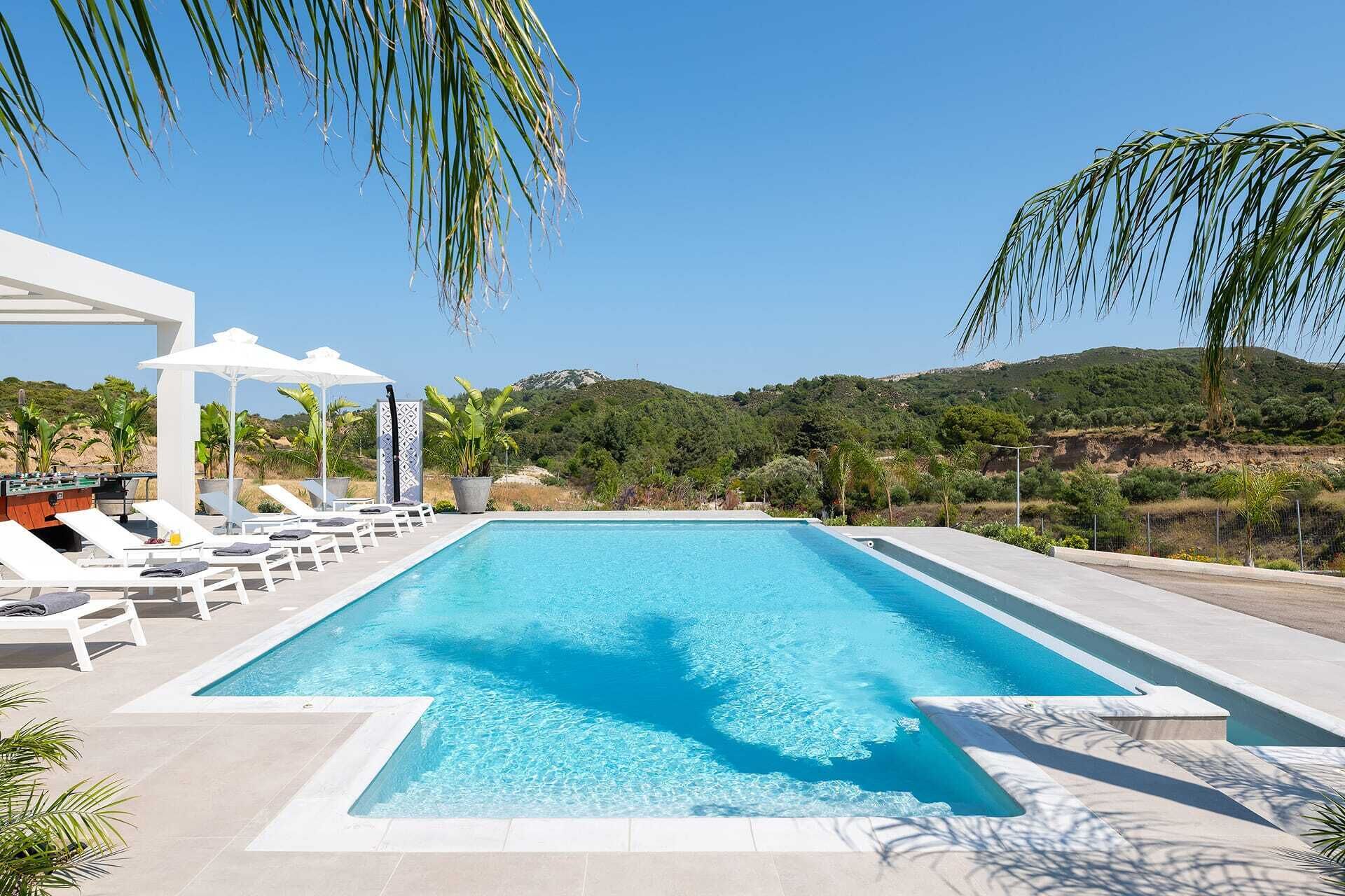 Piscina retangular com água azul cristalina, rodeada por cadeiras de banho brancas e guarda-sóis, com um cenário de vegetação e colinas ao fundo.