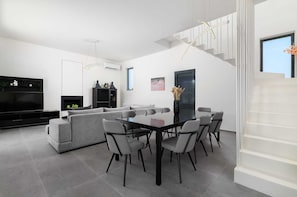 Uma sala de estar moderna com uma TV preta, sofá cinzento e um espaço de jantar com mesa e cadeiras de vidro.