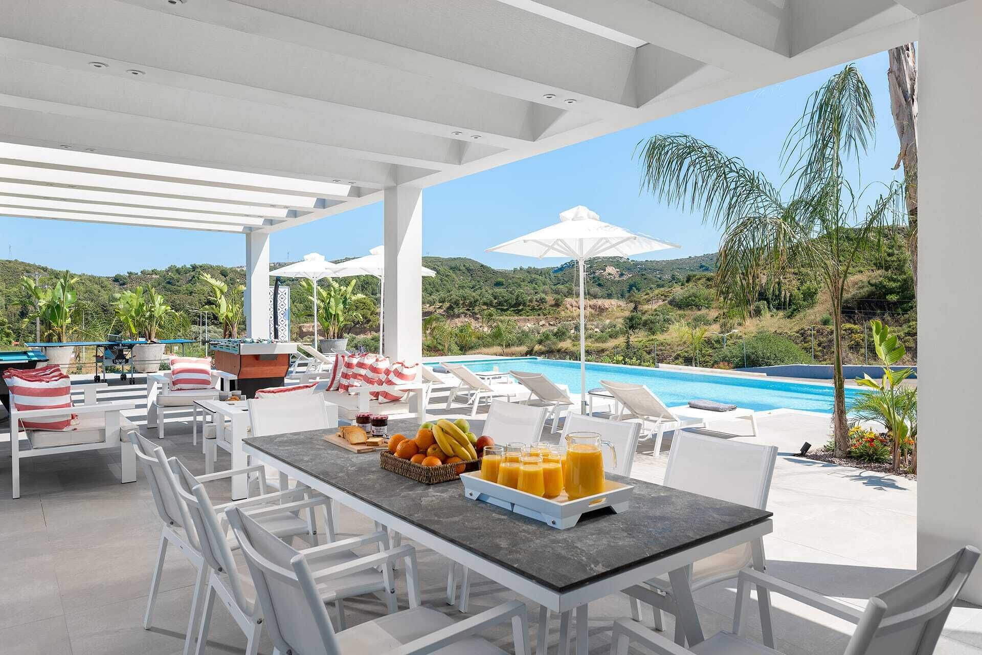 A covered outdoor area with a pool, lounge chairs, and a table with fruit and drinks.