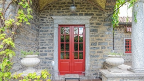 Un bâtiment en pierre avec une porte rouge, deux jardinières en pierre et une petite table.