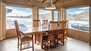 A dining area with a wooden table and chairs, large windows with a view of a lake and mountains, and a hanging pendant light.