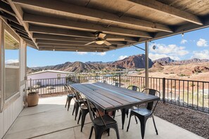 Patio esterno con tavolo e sedie in legno, una ventola a soffitto e vista sulle montagne e su un lago.