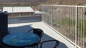 Una terraza en la azotea con una mesa y sillas de vidrio, barandilla blanca y vistas a montañas distantes.