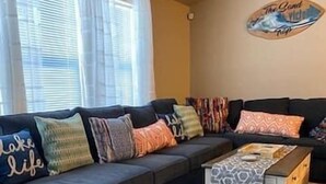 Una sala de estar con un sofá seccional gris oscuro, varios cojines de colores, una mesa de centro estampada y un reloj de pared.