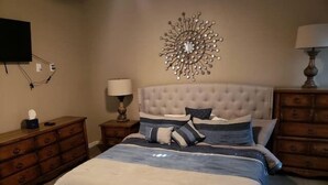 Un dormitorio con cabecera capitonada, cama con colcha gris y azul, dos mesitas de noche con lámparas, cómoda de madera y televisor montado en la pared.