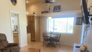 A living room with a sofa, a coffee table, a dining table with chairs, a ceiling fan, and a TV mounted on the wall. - Shoshone One Bedroom with Lake and Pool Views (Lake Havasu City)