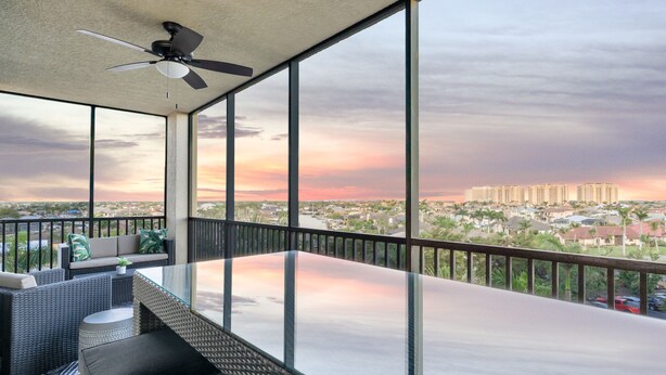 A balcony with a glass table and chairs, a ceiling fan, and a view of a cityscape at sunset. - Condo w/ Views & Resort Amenities in Cape Harbour! (Cape Coral)