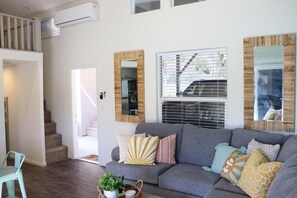 Un salon moderne avec un canapé gris, des coussins décoratifs, une table basse agrémentée d’une plante et un escalier menant à un niveau supérieur.