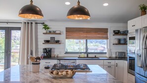 A modern kitchen with a central island, stainless steel appliances, pendant lighting, and a view of the outdoors. - Castle Rock 95, 4brs, Community launch, Riverfront park (Parker)