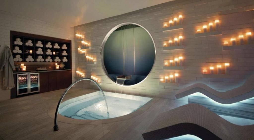 A modern spa room with a circular mirror, a built-in hot tub, and a wall-mounted shelf with decorative items.