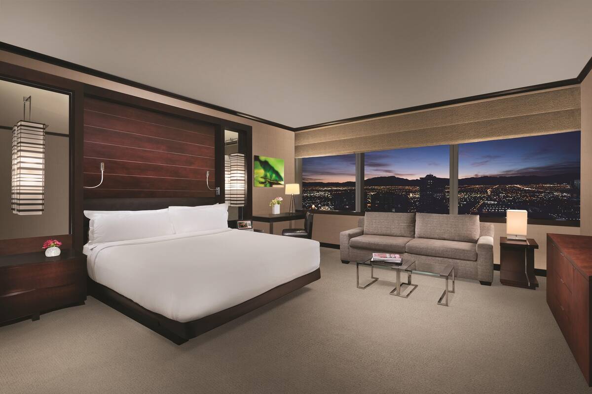 A modern hotel room with a large bed, a sofa, a desk, and a view of a cityscape at night.
