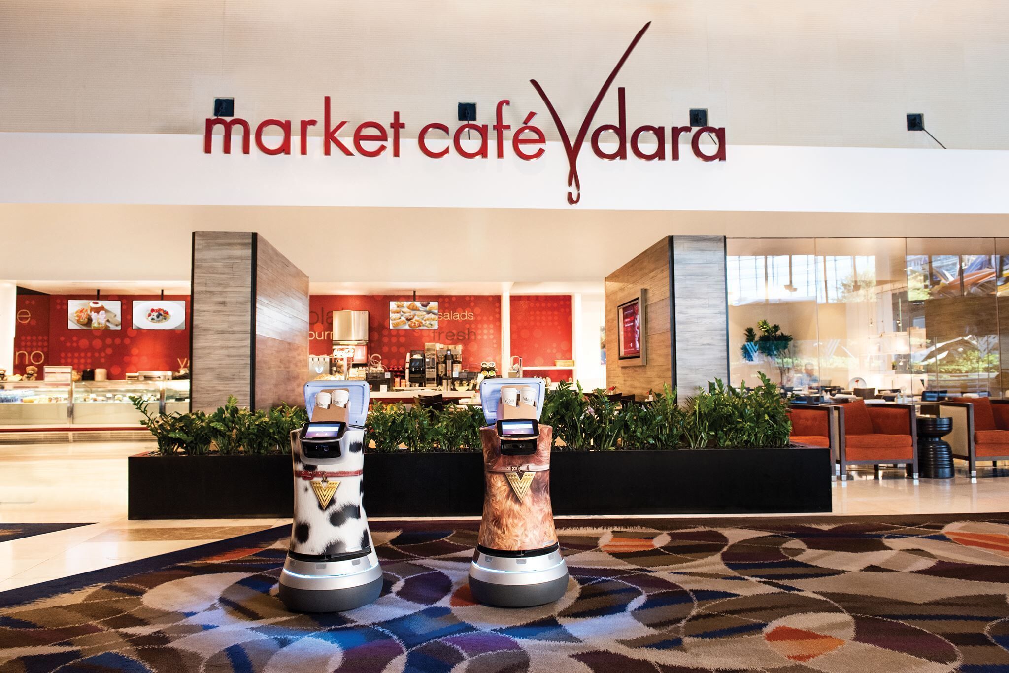 A modern interior with a 'market café' sign, two robotic kiosks, and a seating area with a patterned carpet.