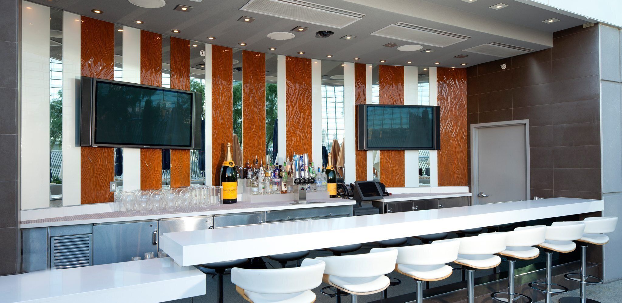 A modern bar area with a long counter, multiple seating options, and two large flat-screen TVs mounted on the wall.