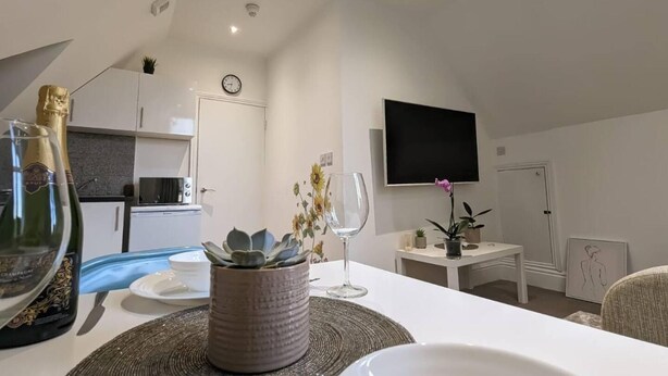 A modern living room with a dining area, a flat-screen TV mounted on the wall, a clock, and a microwave in the kitchen area. - Modern Spacious Apartment Families & Corporate (Hove)
