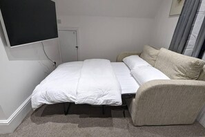 Ein kleines, aufgeräumtes Schlafzimmer mit einem Bett, einer Couch, einem an der Wand befestigten Fernseher und einer Tür.