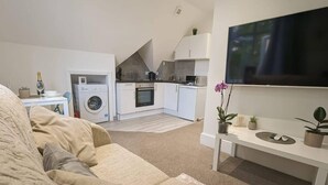 A compact living space with a sofa, a small dining table, a TV, a kitchen area with a washing machine, and a few potted plants. - Modern Spacious Apartment Families & Corporate (Hove)