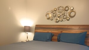 Quarto com cabeceira de madeira, cama com roupa de cama cinza, duas almofadas azuis e um ornamento metálico fixado na parede.