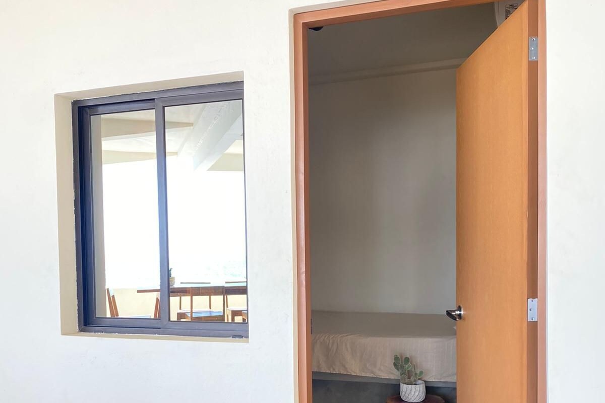 Quarto com uma cama, uma pequena janela e uma porta que dá para outro cômodo.